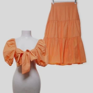 Persikofärgat set med puffärmar - Säljer ett persikofärgat set bestående av en topp med puffiga ärmar och stor knytning framtill samt en matchande volangkjol. Setet är gjort i ett lätt och luftigt material, perfekt för sommarens varma dagar. Toppens rygg har elastisk resår för skön passform.