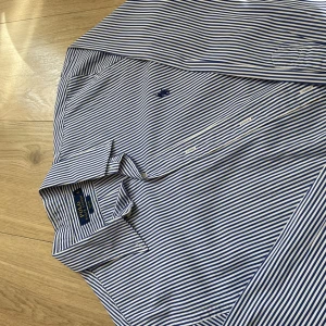 Randig skjorta från Polo Ralph Lauren - Snygg blåvit randig skjorta från Polo Ralph Lauren i slim fit-modell. Skjortan har klassisk krage, långa ärmar och knäppning framtill. Tillverkad i bomull och prydd med den ikoniska broderade loggan på bröstet. Perfekt för en clean och stilren look. Helt ny.