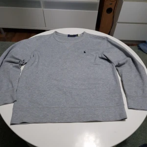 Grå sweatshirt från Polo Ralph Lauren - Klassisk grå sweatshirt från Polo Ralph Lauren med rund halsringning och diskret broderad logga på bröstet. Tröjan har lång ärm och är tillverkad i mjuk bomull för en bekväm känsla. Perfekt till en avslappnad stil.