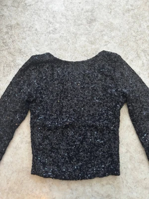 Svart glittrig långärmad topp - Svart långärmad topp med glittriga paljetter över hela ytan. Toppen har en rund halsringning och är croppad. Materialet är troligtvis polyester och syntet, vilket ger en stretchig och bekväm passform. Perfekt för dig som vill sticka ut med lite extra glitter.