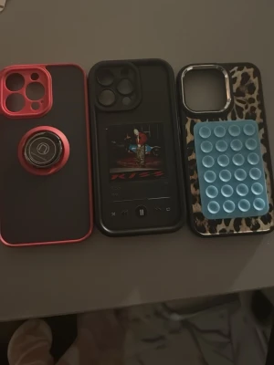 3 unika mobilskal till iPhone - Säljer tre olika mobilskal till iPhone. Ett svart och rött skal med metallring för magnetfäste, ett svart skal med tryck av en musikspelare och ett leopardmönstrat skal med blå pop it-detalj. Alla skal är i plast och har öppningar för kamera och knappar. Och screen protection