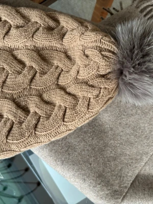 Beige kabelstickad mössa med pälstofs från S’Max Mara - Säljer en beige kabelstickad mössa med stor, fluffig pälstofs i grått. Mössan har ett snyggt flätmönster och är gjord i 80 % ull och 20 % cashmere. Helt ny och oanvänd. 