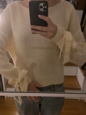 Krämvit stickad tröja med knytbar rosett - Superfin krämvit stickad tröja från Meet Me There. Tröjan har långa ärmar med snygg knytbar rosett vid ärmslutet och ribbade muddar. Aldrig använd 