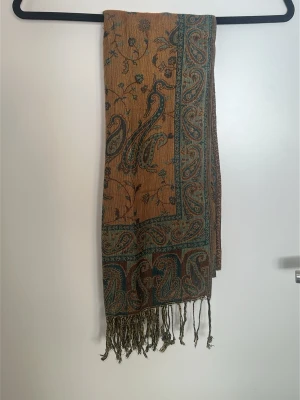 Brun paisleymönstrad halsduk - Säljer en brun halsduk med paisleymönster i grönt och vinrött. Halsduken har fransar i ändarna och ett detaljerat, orientaliskt inspirerat tryck. Perfekt accessoar för att lyfta din outfit med lite bohemisk känsla.