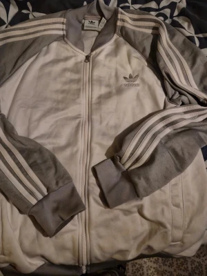 Gråvit Adidas zip-tröja med logga XL - Säljer en gråvit zip-tröja från Adidas med klassiska tre ränder längs ärmarna och broderad logga på bröstet. Tröjan har ribbade muddar och dragkedja framtill. Perfekt för en sportig och avslappnad look. Storlek XL