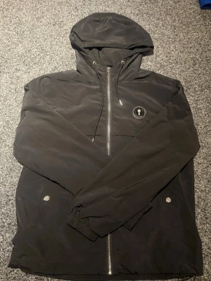 Svart vindjacka från Trapstar - Snygg svart vindjacka från Trapstar med huva och dragkedja framtill. Jackan har två fickor med tryckknappar, elastiska muddar vid ärmsluten och Trapstar-logga på bröstet samt stor text på ryggen. Perfekt för streetwear-stilen.