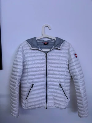 Colmar Jacka - Snygg vit pufferjacka från Colmar med grått innerfoder och huva. Jackan har två dragkedjeförsedda fickor framtill och en logga på ärmen. Lättviktigt material och quiltad design gör den perfekt för kalla dagar.