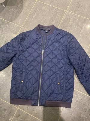 Ralp Lauren Jacka  - Snygg marinblå bomberjacka från Polo Ralph Lauren med quiltat mönster, ribbade muddar och krage i mörkare ton. Jackan har dragkedja framtill, två fickor med tryckknappar och diskret broderad logga på bröstet. Perfekt för dig som vill ha en stilren och trendig look. Lite solblekt i kragen. Motsvara ungefär S. 