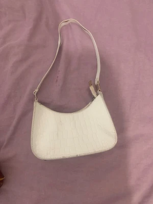 Vit axelväska med krokopräglat mönster - Snygg vit axelväska i konstläder med krokopräglat mönster och guldfärgade detaljer. Väskan har en enkel, stilren form och justerbar axelrem. Perfekt för dig som gillar trendiga accessoarer och vill ha en väska som sticker ut.