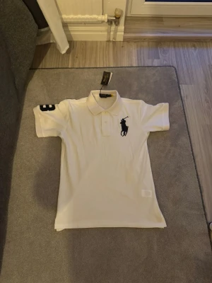 Vit pikétröja från Polo Ralph Lauren - Klassisk vit pikétröja från Polo Ralph Lauren med stor mörkblå logga på bröstet och siffran 3 på ärmen. Tröjan har krage, knappar framtill och korta ärmar. Perfekt för en clean och sportig stil.