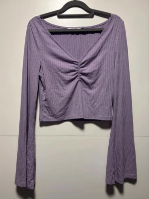 Ljuslila ribbad croppad topp - Trendig ljuslila croppad topp med lång ärm och v-ringning. Toppen har ribbad struktur och snygg rynkning framtill som ger en cool detalj. Perfekt att styla med höga jeans eller kjol. Från NA-KD i storlek S