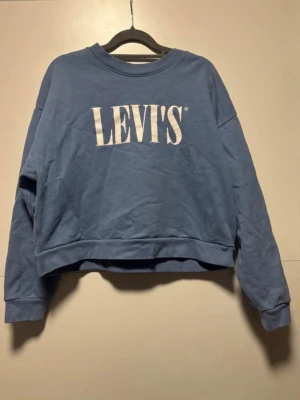Blå cropped Levi's hoodie - Säljer en blå cropped hoodie från Levi's med stort vitt tryck på bröstet. Tröjan har rund halsringning, långa ärmar och en avslappnad passform. Perfekt för dig som gillar streetwear och vill ha något bekvämt och stilrent.