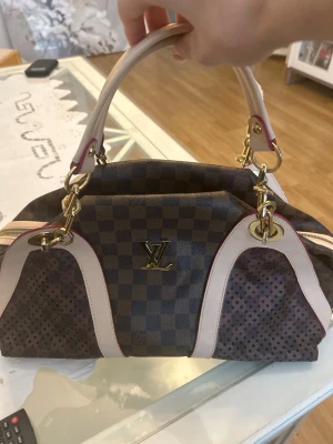 Louis Vuitton brun handväska (Äkta) - Snygg handväska från Louis Vuitton i klassiskt brunt damier-mönster med ljusa läderdetaljer och guldfärgade metalldelar. Väskan har två handtag och dragkedja upptill. Insidan är fodrad i starkt rosa tyg. Perfekt för dig som vill ha en lyxig accessoar.