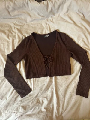 Brun croppad ribbad topp H&M - Säljer en brun croppad topp från H&M med långa ärmar och v-ringning. Toppen har ribbad struktur och en söt knytning framtill. Perfekt att styla med höga jeans eller kjol för en trendig look.