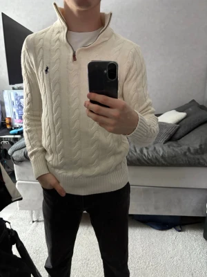 Vit kabelstickad tröja Polo Ralph Lauren - Kabelstickad vit tröja från Polo Ralph Lauren med halv dragkedja och hög krage. Tröjan har det klassiska mörkblå logobroderiet på bröstet och ribbade muddar vid ärmslut och nederkant. Perfekt för en clean och stilren look.