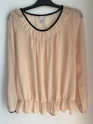 Ljusrosa blus med volangkant från VILA - Söt ljusrosa/ ljusaprikos blus från VILA med rundad halsringning och svarta detaljer. Blusen har långärmad design, lätt transparent material och volangkant nedtill. Perfekt för en trendig och feminin look.