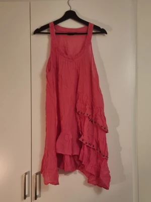 Rosa asymmetrisk klänning Lindex - Säljer en rosa klänning från Lindex i storlek L. Klänningen har asymmetrisk nederkant, volangdetaljer och dekorativa knappar på framsidan. Ärmlös modell med breda axelband och v-ringad hals. Perfekt för sommarens alla tillfällen!