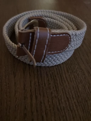 Beige flätat bälte med skinn-detaljer - Snyggt beige flätat bälte med bruna skinn-detaljer och metallspänne. Perfekt accessoar för att lyfta din outfit med en stilren touch. Passar till både jeans och chinos.