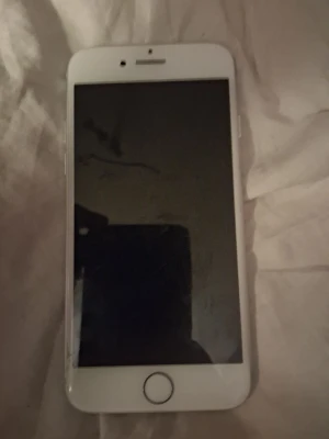 Vit iPhone 7 - Säljer en vit iPhone med klassisk rund hemknapp och stor skärm. Telefonen har en stilren design i aluminium och glas, med framsida i svart och ram i vitt. Perfekt för dig som gillar Apples enkla och snygga look.