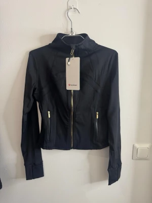 Svart träningsjacka från Lululemon storlek 6 - Snygg svart träningsjacka från Lululemon med guldfärgade dragkedjor och hög krage. Jackan har två fickor med dragkedja och är tillverkad i ett stretchigt funktionsmaterial som passar perfekt till träning eller som ett sportigt lager.