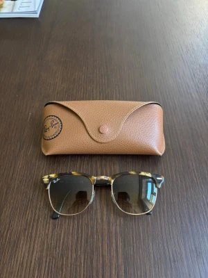Ray-Ban Clubmaster solglasögon - Snygga Ray-Ban Clubmaster solglasögon med klassisk brunmelerad båge och guldfärgade detaljer. Glasen är tonade i brunt och formen är retroinspirerad med markerad ovandel. Medföljer original Ray-Ban fodral i brunt skinn.