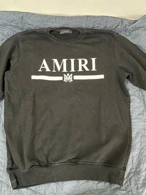 Svart sweatshirt från Amiri - Säljer en svart sweatshirt från Amiri med stor vit logga och grafisk detalj på bröstet. Tröjan har rund halsringning, långa ärmar och ribbade muddar vid ärmslut och nederkant. Tillverkad i mjukt bomullsmaterial som är skönt att bära.