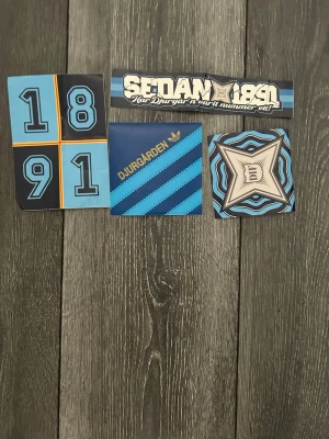 Djurgården klistermärken samling - Säljer ett set med fyra olika Djurgården-klistermärken i blå, svart och guld. Motiven inkluderar årtalet 1891, texten 'Sedan 1891', Djurgården-logga och Adidas-inspirerad design. Perfekt för att dekorera laptop, mobil eller anteckningsbok.