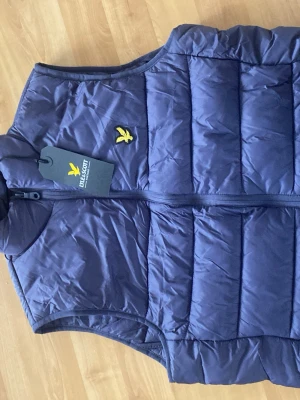  Helt ny Mörkblå dunväst från Lyle & Scott - Snygg mörkblå dunväst från Lyle & Scott med gul logga på bröstet. Västen har quiltad design, dragkedja framtill och är tillverkad i ett lätt, vadderat material som håller värmen. Perfekt att slänga över en hoodie eller tröja för extra stilpoäng.                                     Har inte använd den någonting säljer pga den inte passade tog fel storlek. Ny pris på denna är 1200kr.