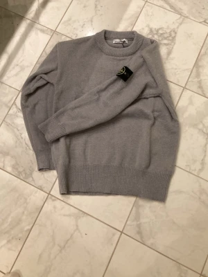 Ljusgrå tröja från Stone Island - Säljer en stilren ljusgrå tröja från Stone Island med klassisk rund halsringning och ribbade muddar. Tröjan har den ikoniska Stone Island-loggan på ärmen och är tillverkad i mjukt stickat material. Perfekt för dig som gillar streetwear och enkel design.