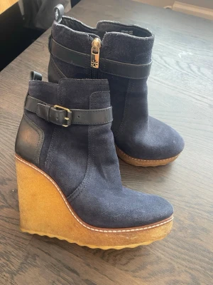 Kilklackade stövletter från Tory Burch - Mörkblå stövletter i mocka från Tory Burch med hög kilklack i naturfärgat gummi. Dekorativt spänne och dragkedja med logga i guld. Snygg kontrastsöm längs sulan och detaljer i svart läder. Perfekta för dig som vill ha både höjd och stil.