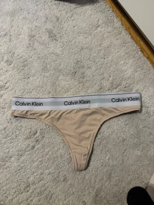 Beige stringtrosa från Calvin Klein - Snygg beige stringtrosa från Calvin Klein med bred resår i midjan och logga runtom. Trosan har en klassisk stringmodell och är tillverkad i mjukt och stretchigt material för en bekväm passform.