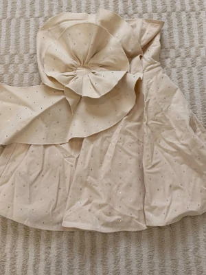 Krämvit/beige klänning med stor rosett - Supersnygg kort klänning med en stor dekorativ rosett framtill. Klänningen är täckt av små glittriga silver detaljer och har en elegant, axelbandslös design. Perfekt för dig som vill sticka ut med en unik look. Den sitter uppe väldigt bra då det finns en remsa som gör att den håller sig på sin plats. Köpt för en månad sedan för 2000kr på asos hemsida, endast använd 1 gång - skriv för bilder på