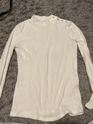 Vit ribbad långärmad topp från Mango - Säljer en vit ribbad långärmad topp från Mango med snygga guldfärgade knappar på axeln. Toppen har en ganska hög hals. den är knappt använd o är jätte fin för en classy look