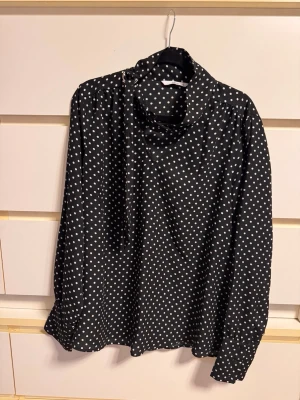 Svart prickig blus med knytkrage - Snygg svart blus med vita prickar och trendig knytkrage. Blusen har långa ärmar och en lös, avslappnad passform. Perfekt för dig som gillar klassiska mönster med en twist. Materialet känns mjukt och följsamt.