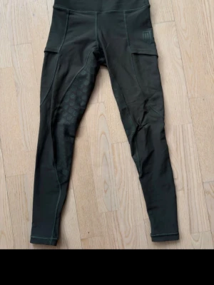 Mörkgröna ridbyxor från Maya Delorez - Snygga mörkgröna sportleggings från Maya Delorez med hög midja och smart ficka på låret. Materialet är stretchigt och har ventilerande partier med hexagonmönster på insidan av benen. Perfekta för träning eller ridning. Hela och fina! 2XS 