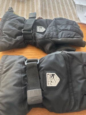 Svarta vinterhandskar från Hestra - Svarta vinterhandskar från Hestra med justerbart spänne vid handleden och klassisk logga på ovansidan. Handskarna är tillverkade i slitstarkt syntetmaterial och har en längre mudd för extra skydd mot kyla. Perfekta för kalla dagar utomhus.