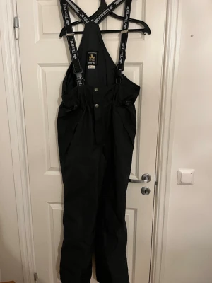 Svarta täckbyxor Terinit Gore-Tex - Svarta täckbyxor från Terinit med Gore-Tex-material och justerbara hängslen. Byxorna har hög midja, knäppning framtill och är perfekta för kalla dagar. Sportig design med logotyp på hängslena och insidan. Grymma för vinteraktiviteter.