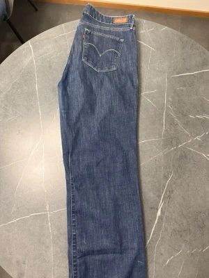 Blå raka jeans från Levi's - Säljer ett par klassiska blå Levi's Curve Classic Straight jeans med raka ben och normalhög midja. Jeansen har fem fickor, guldfärgade detaljer och Levi's ikoniska bakficka med söm. Perfekta till en avslappnad stil. (Framfickorna är sydda typ ish stängda)