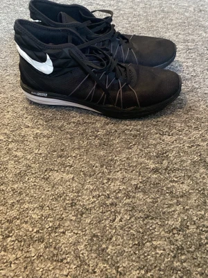 Svarta Nike midsneakers mesh - Säljer ett par svarta Nike sneakers med vit swoosh på sidan och meshmaterial för extra andningsförmåga, perfekta för gymmet. Skorna har svarta snören, diskret logga på plösen och en grå sula med texten Dual Fusion. Snygg design med tunna linjer längs sidorna. Knappt använda. Priset är ej hugget i sten 