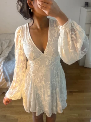 Vit blommig kortklänning med volang - Superfin vit kortklänning med blommigt mönster och volangdetaljer. Klänningen har v-ringning, långa puffiga ärmar och öppen rygg. Perfekt för sommarens alla tillfällen och ger en romantisk vibe. Passar jätte bra för er som är mellan storlek 34-38 ( knytet i ryggen gör storleken väldigt skysterbar) 