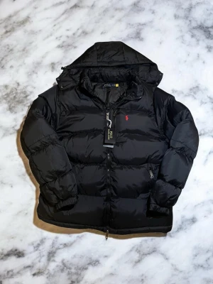 Svart pufferjacka från Polo Ralph Lauren - Snygg svart pufferjacka från Polo Ralph Lauren med huva och dragkedja. Jackan har det klassiska röda Polo-logotypen broderad på bröstet och två sidofickor med dragkedja. Perfekt för kalla vinterdagar och riktigt stilren look. ⚠️ Är stor i storlek, passar mer M och L ⚠️ 