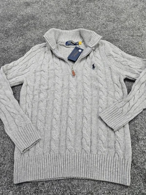 Grå kabelstickad tröja Polo Ralph Lauren - Snygg grå kabelstickad tröja från Polo Ralph Lauren med halv dragkedja och hög krage. Tröjan har ribbade muddar och nederkant samt den klassiska mörkblå loggan broderad på bröstet. Perfekt för lager på lager och stilren look.