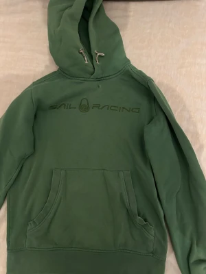 Grön hoodie från Sail Racing - Snygg grön hoodie från Sail Racing med stor ficka framtill och justerbar huva med snören. Loggan är broderad på bröstet i matchande grön färg. Hoodien har ribbade muddar vid ärmslut och nederkant. Perfekt för en avslappnad och sportig stil.