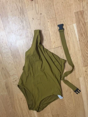 Olivgrön baddräkt oneshoulder - Snygg olivgrön baddräkt med oneshoulder-design och matchande bälte med spänne. Perfekt för dig som vill sticka ut på stranden med en modern och trendig look.