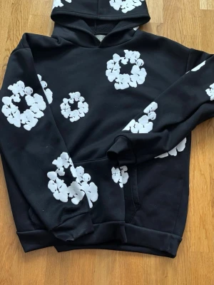 Svart hoodie från Denim Tears  - Svart hoodie från Denim Tears med vita blomliknande tryck över hela plagget. Klassisk känguruficka framtill och ribbade muddar vid ärmar och nederkant. Tillverkad i mjukt material och har en stor huva för extra komfort.