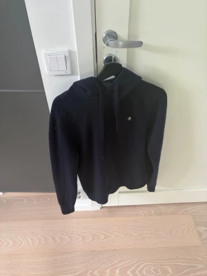 Mörkblå hoodie med broderad logga - Mysig mörkblå hoodie med huva och dragsnören. Tröjan har en liten broderad logga på bröstet och är tillverkad i ett mjukt, stickat material. Perfekt för dig som gillar en enkel men stilren look.