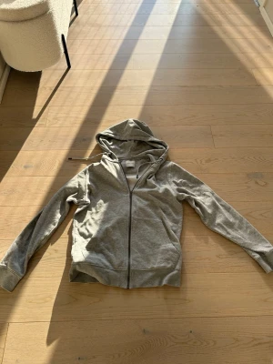 Grå zip hoodie från Jack & Jones med huva - En enkel och stilren grå hoodie med dragkedja framtill och huva. Hoodien har långa ärmar, ribbade muddar vid ärmslut och nederkant samt två fickor framtill. Perfekt för en avslappnad och chill look.