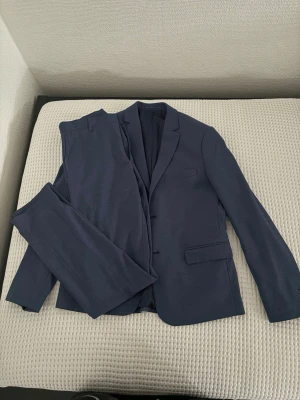 Mörkblå kostym med kavaj och byxor - Snygg mörkblå kostym med klassisk kavaj och matchande byxor. Kavajen har två knappar, smala slag och fickor med lock. Perfekt för dig som vill ha en stilren och tidlös look. Materialet känns lätt och passar året runt.