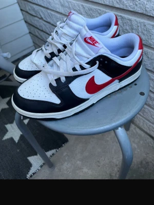 Nike Dunk Low svart/vit/röd - Säljer ett par Nike Dunk Low sneakers i svart, vitt och rött. Skorna har klassisk låg siluett, vit ovandel i läder med svarta overlays och röd Swoosh samt häl. Tjock vadderad plös och vita skosnören. Perfekta för dig som gillar streetwear och vill sticka ut.
