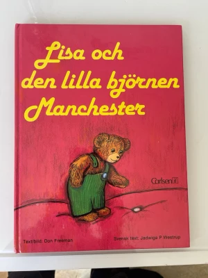Lisa och den lilla björnen Manchester - Barnbok med hårda pärmar, illustrerad i färg. Framsidan visar en nallebjörn i gröna byxor mot en rosa bakgrund. Boken är skriven av Don Freeman med svensk text av Jadwiga P Westrup. Perfekt för dig som gillar klassiska berättelser och fina illustrationer.